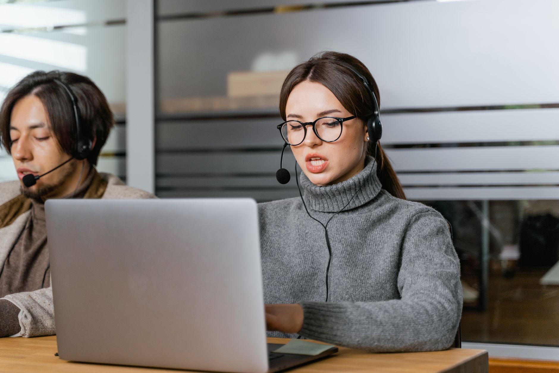 Una donna con occhiali, cuffie e microfono parla mentre usa un portatile; alle sue spalle un collega con le cuffie
